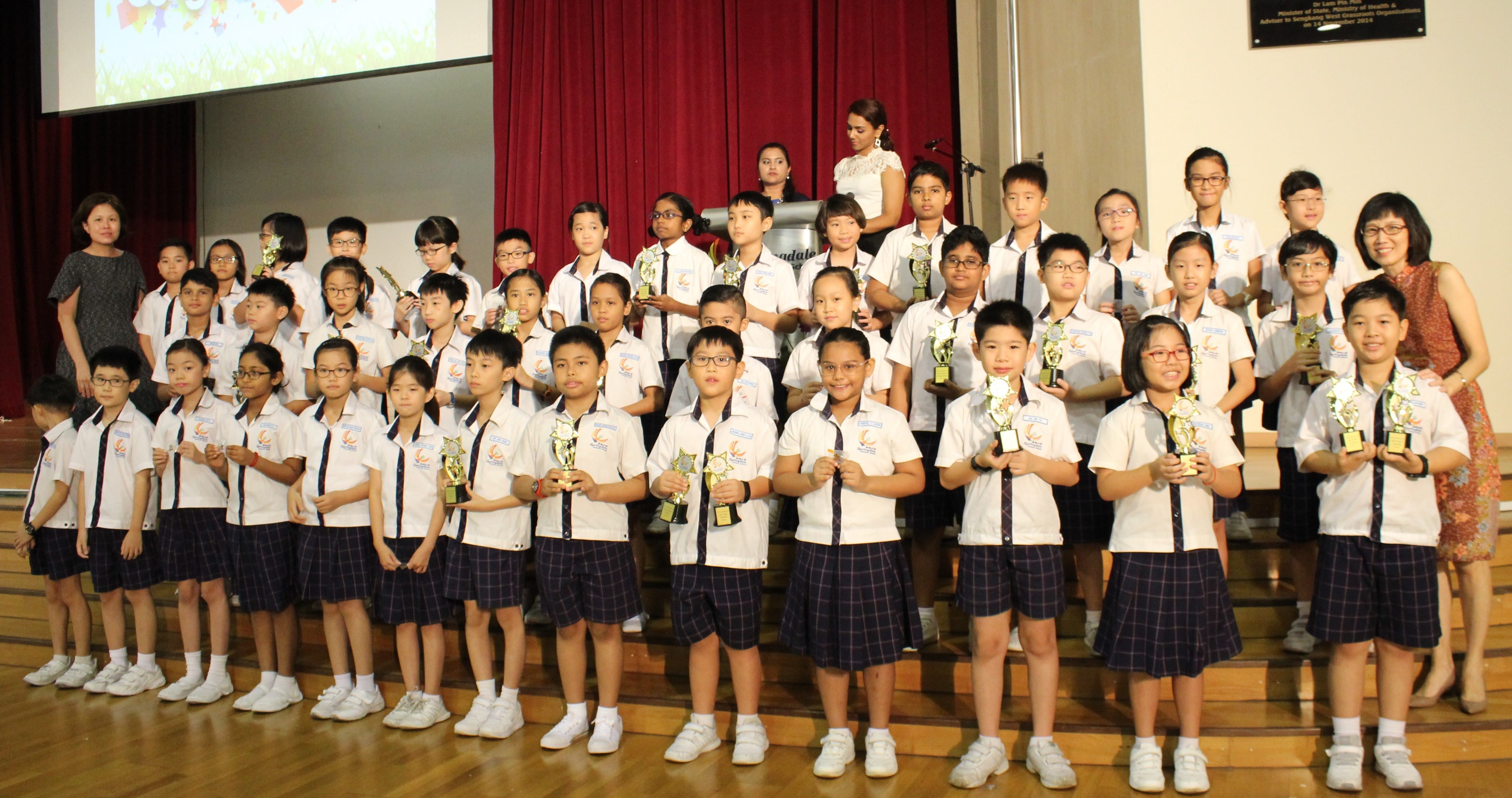 Large group of school children in white uniforms holding trophies on a stage with teachers nearby.
